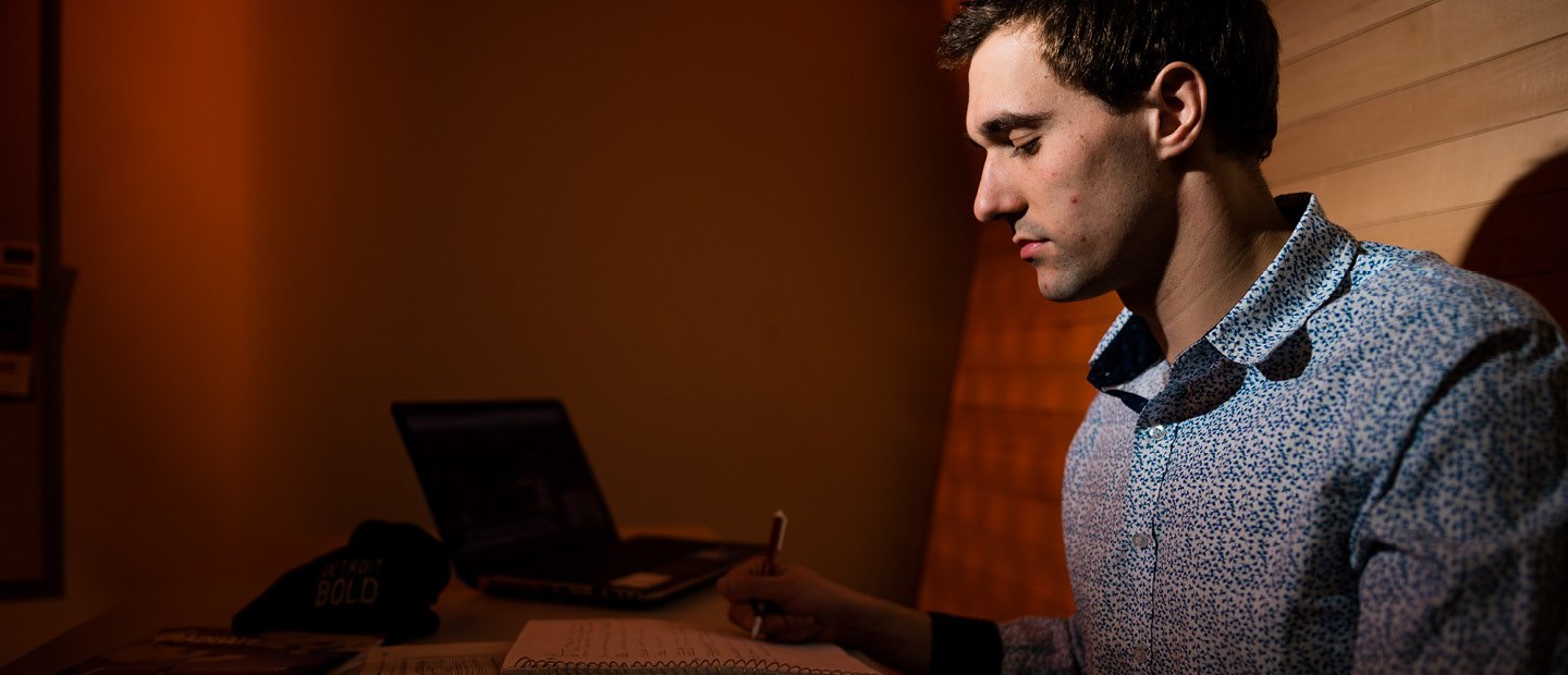 一个男人坐在一个展位,写在一本笔记本和一个开放的笔记本电脑在他旁边。