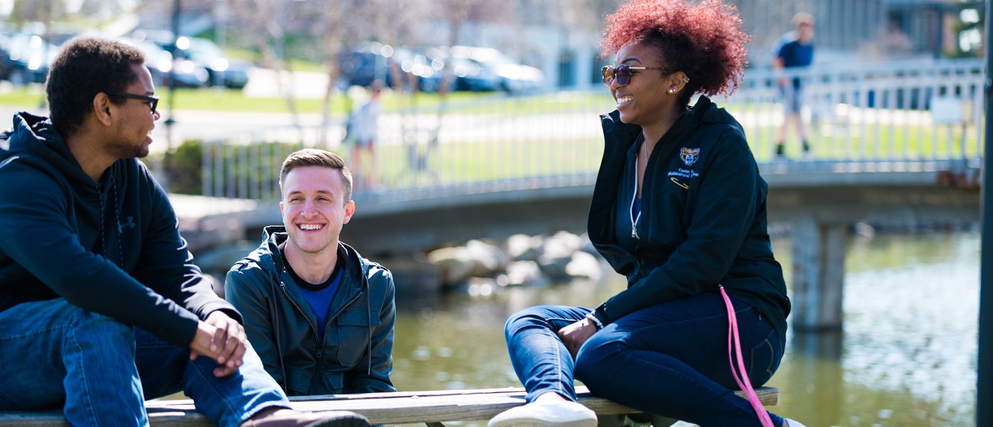 两位男学生和一位女学生坐在外面的湖前，背景是一座桥