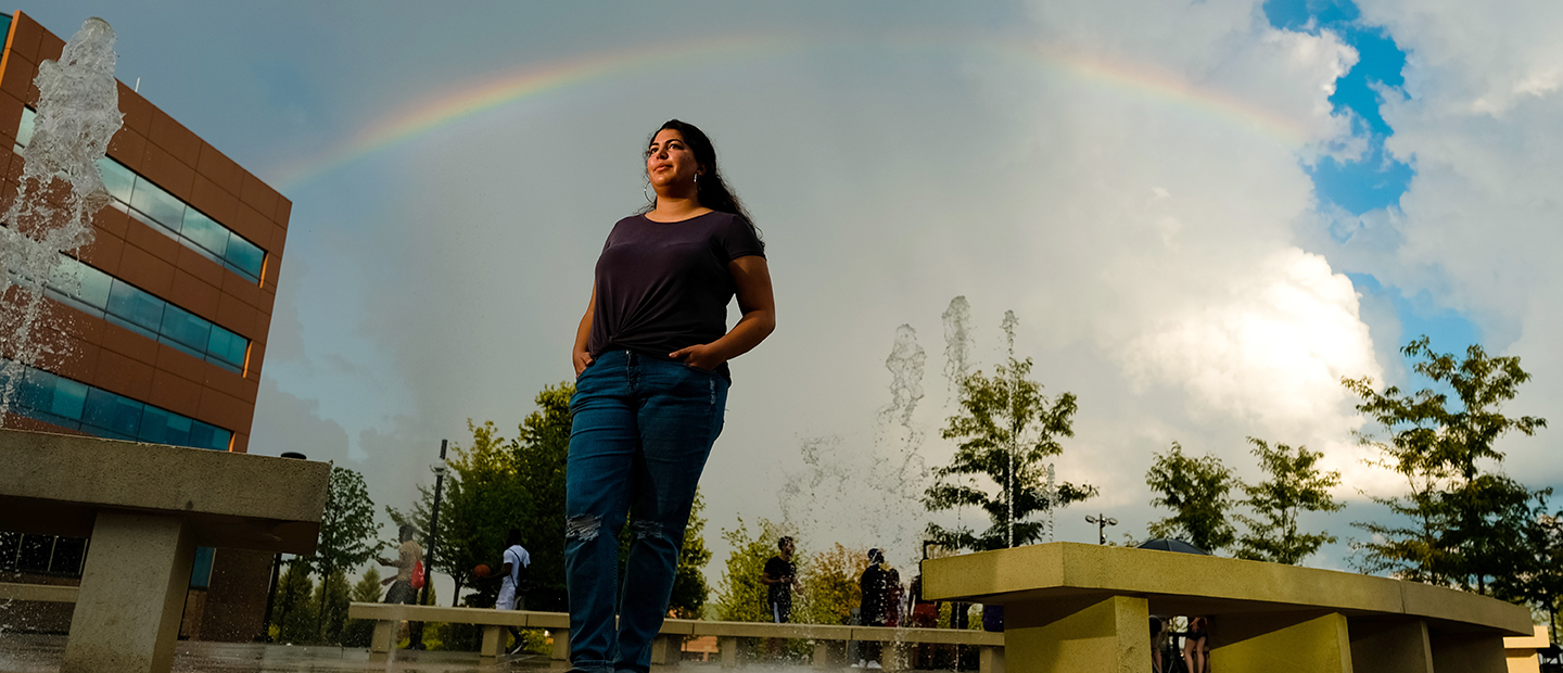 一个年轻的女人站在她的手在她的口袋在奥克兰大学的校园,彩虹和喷泉。万博ManBetX登录