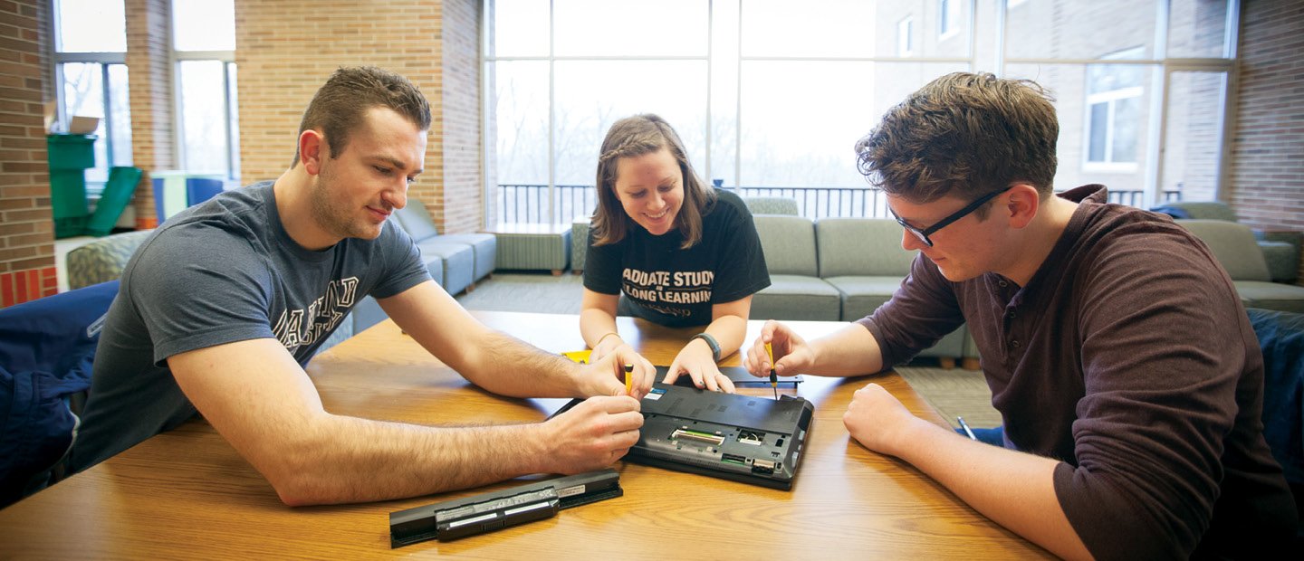 三个学生围坐在桌子旁，用螺丝刀检查一台笔记本电脑