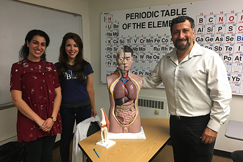教授站在元素周期表的图表前，拿着一个人体躯干模型，两个学生在他的右边