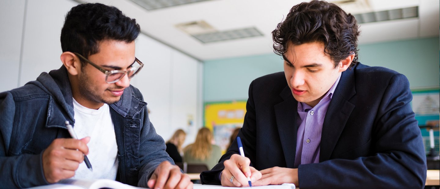 两个年轻人坐在教室的桌子前，在笔记本上写字