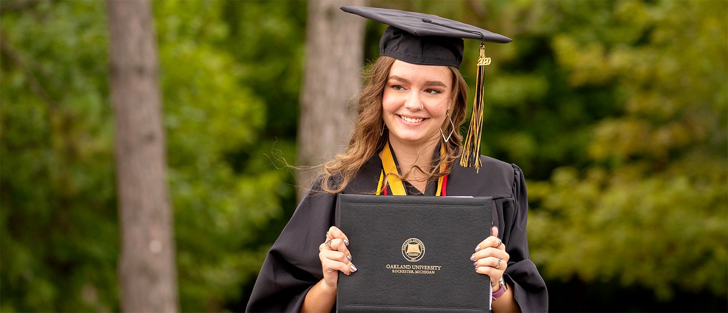 一位奥克兰大学的女万博ManBetX登录毕业生，戴着帽子，穿着长袍，手里拿着文凭。