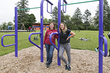 图片中，两个女孩倚靠在公园的设施上，背景是秋千和自行车