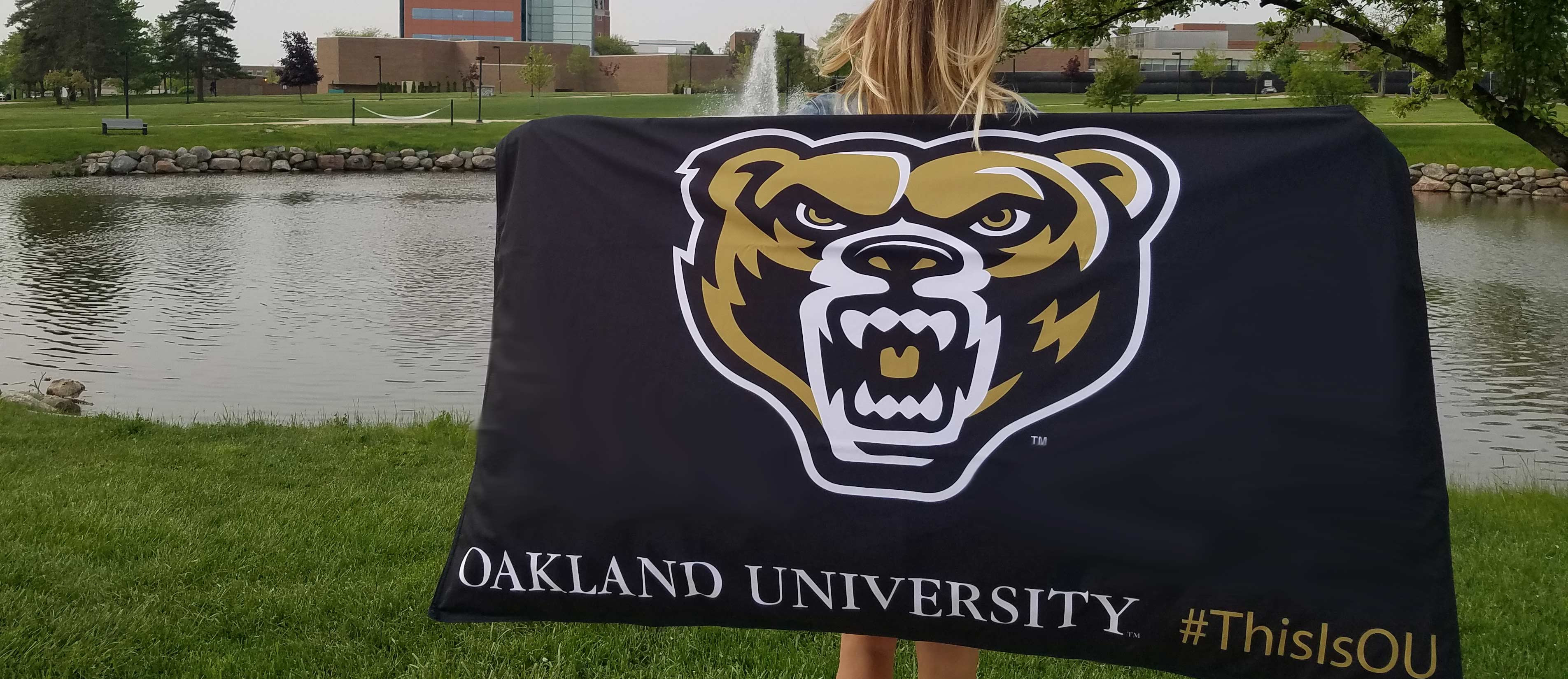 女人拿着5英尺3英尺的国旗在熊湖的前面。国旗有熊头和奥克兰大学# ThisIsOU印刷。万博ManBetX登录
