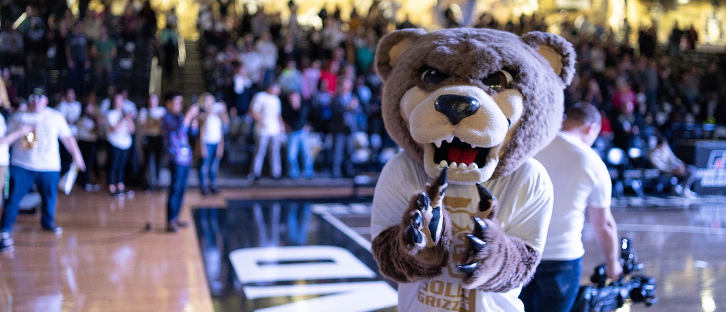 Grizz bear mascot standing on an athletic court with people seated in the stands