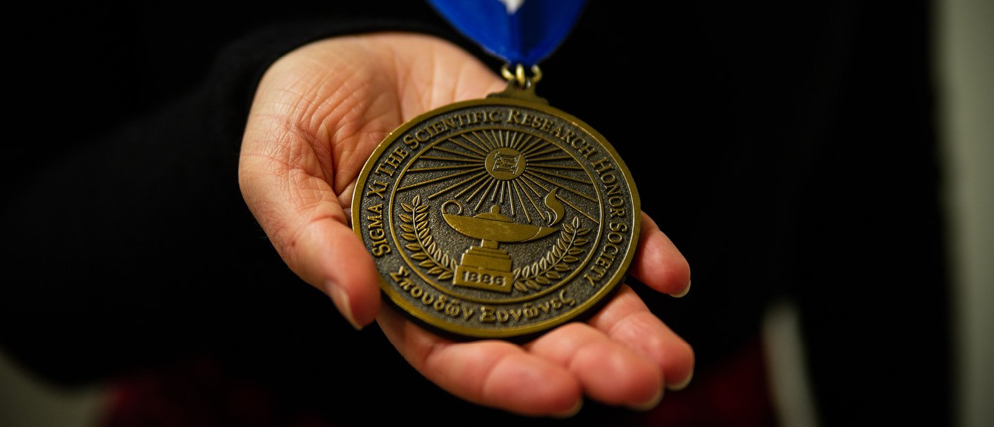 一只手拿着一枚金牌。文本奖章说:“Sigma Xi科研荣誉社会”