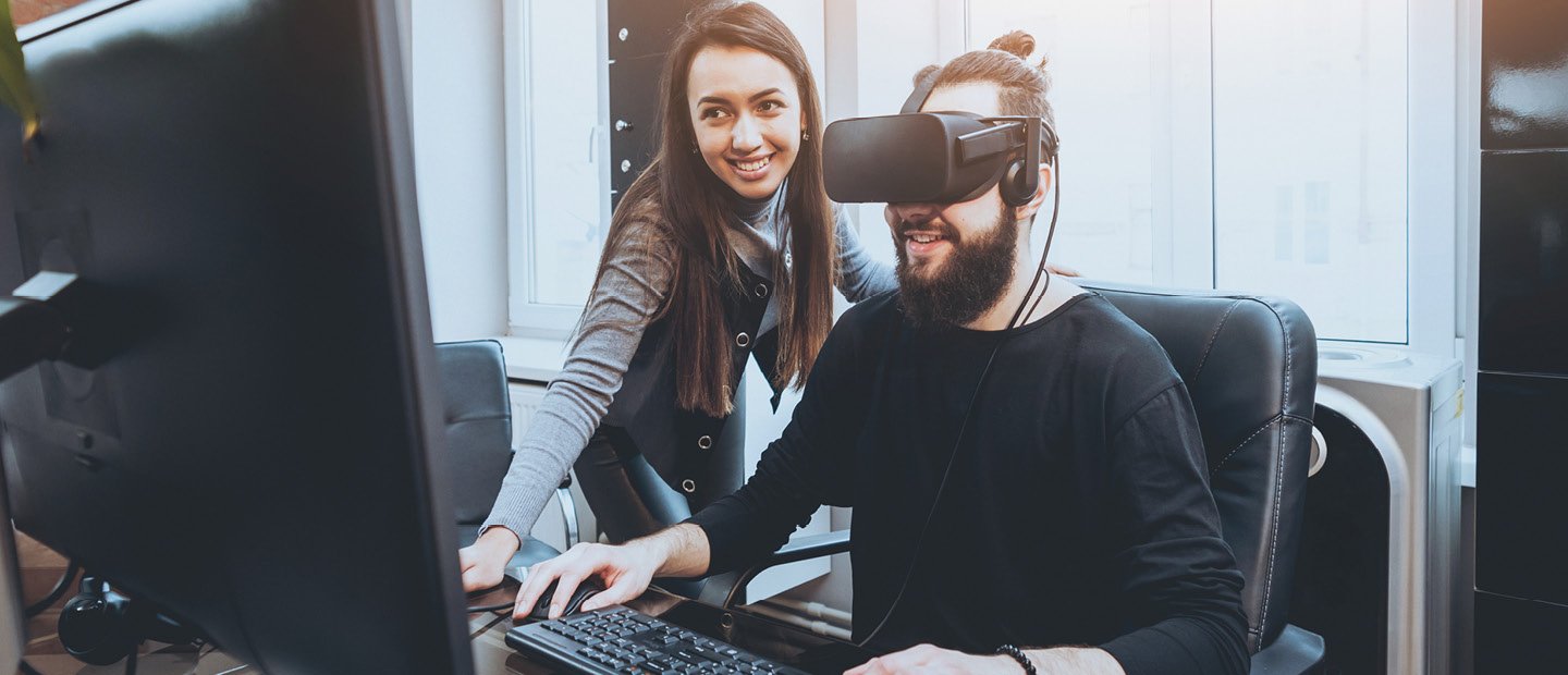 男人穿一件黑色虚拟现实耳机,坐在一台电脑和一个女人站在他旁边