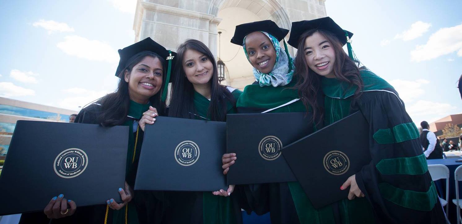 四名毕业生手持毕业证，头戴学士帽，身穿学士服，站在艾略特塔前。