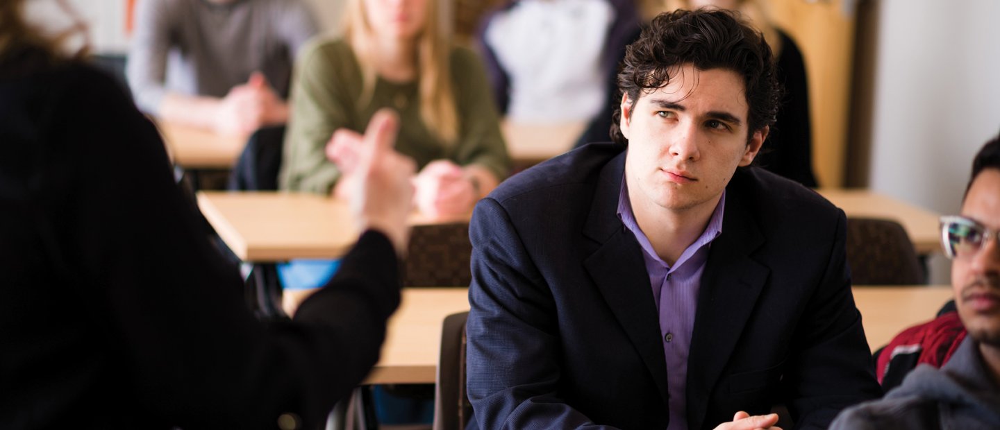 一个年轻人在一个满是学生的教室里，看着教室前面的一个人